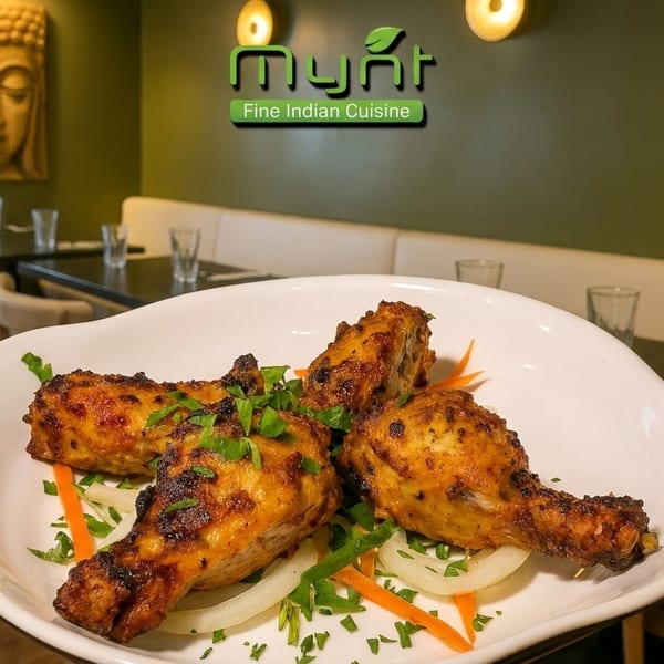 Four chicken drumsticks on a white plate with onions and herbs, in a restaurant setting.