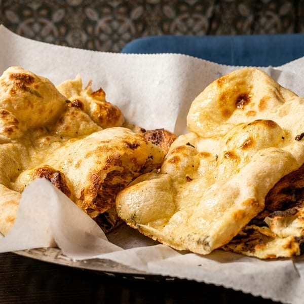 A basket of warm, golden-brown naan bread, some with charred spots, lined with white paper.